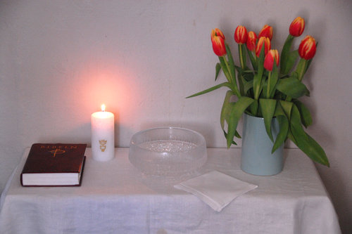 Kastepöytä jossa kynttilä, kastemalja, raamattu ja punaisia tulppaaneja